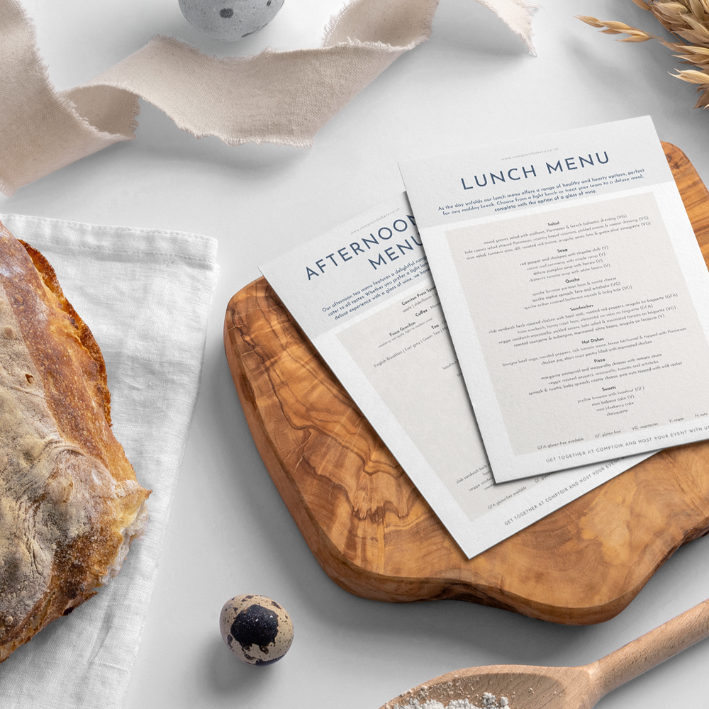 Comptoir Bakery catering menus displayed on a wooden board with a rustic bread and wooden spoon in the foreground.