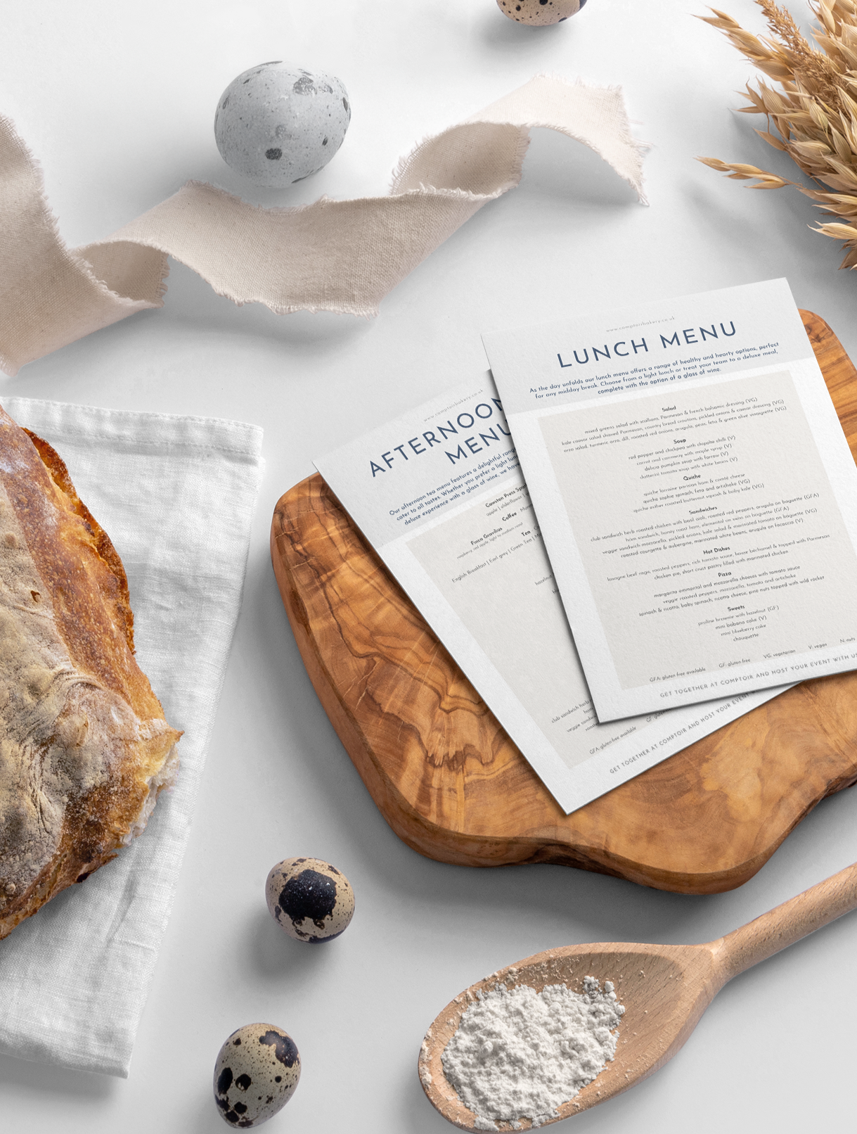 Comptoir Bakery catering menus displayed on a wooden board with a rustic bread and wooden spoon in the foreground.