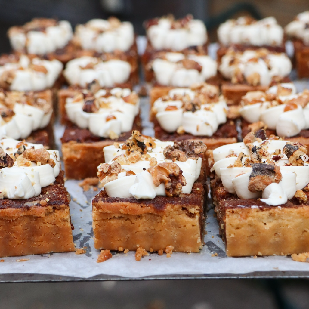 Square pecan caramel bars topped with cream and candied nuts at Comptoir Bakery, freshly prepared on a baking tray.