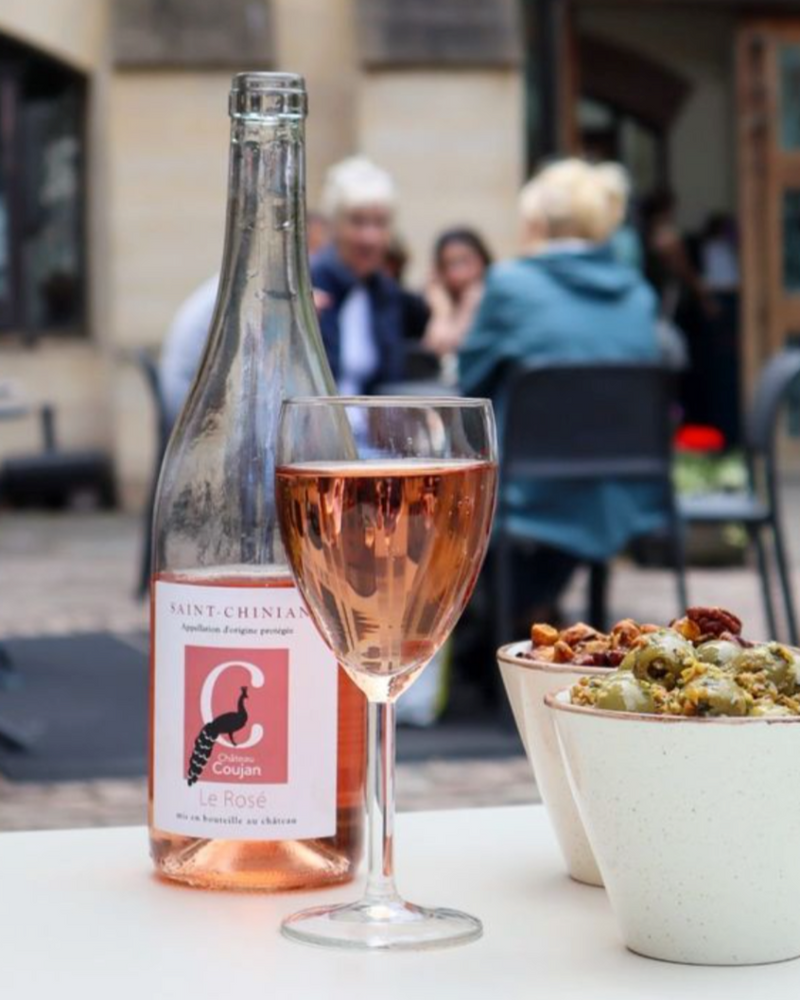 Glass of rosé wine with a bottle of Château Coujan Le Rosé and bowls of marinated olives and nuts at an outdoor seating area at Comptoir Bakery.